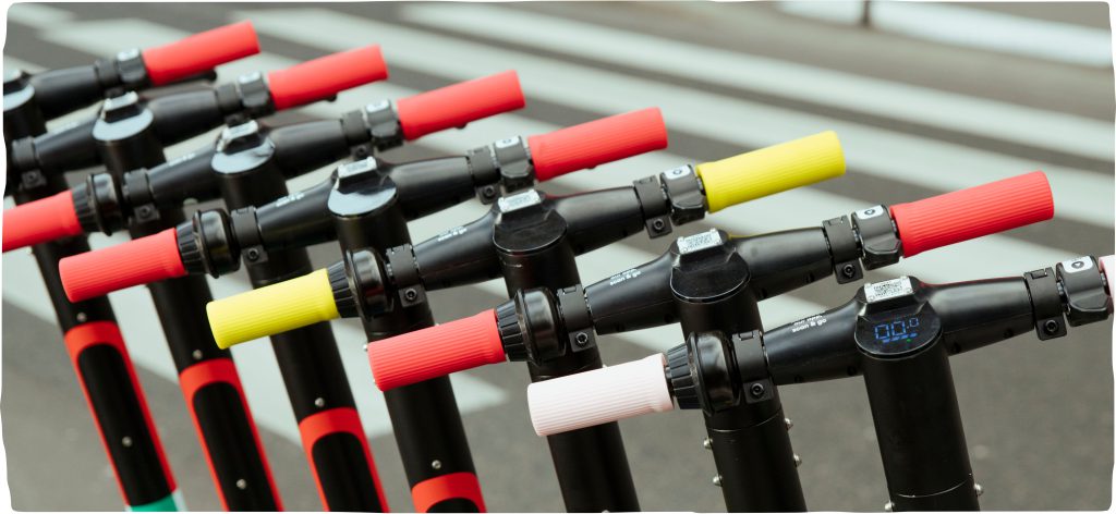 An image of the red, yellow and light-pink handlebars of seven Dott scooters lined up neatly in a row.