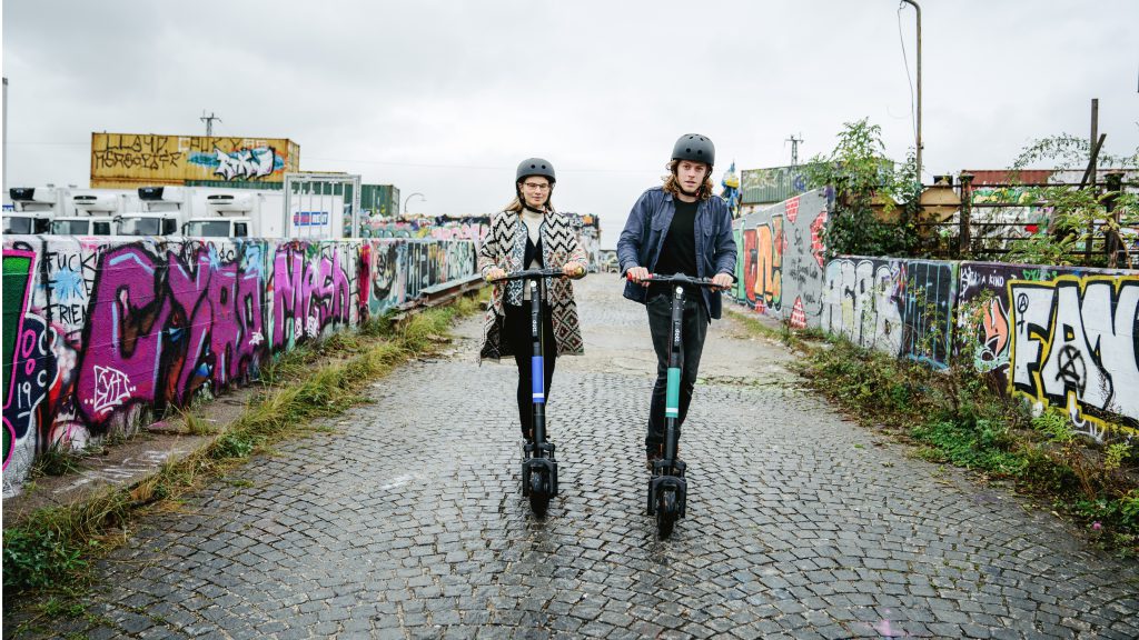 Two young adults wearing helmets are riding Dotts towards the camera. The graffiti in the background brings color to the contrasting gray sky.