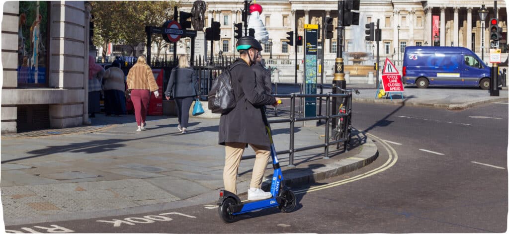 A commuting man stops at traffic lights. He wears a backpack and has one foot on his Dott and one foot on the road. The Charing Cross Underground station is visible in the background.